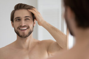 Happy handsome young guy combing smooth brown hair with fingers at mirror in bathroom, looking at reflection with toothy smile. Man satisfied with haircare cosmetic product, enjoying bath routine