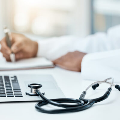 Laptop, stethoscope and doctor writing in notebook for research planning or medical tech innovation in hospital office. Healthcare medic worker, research strategy book notes and online communication